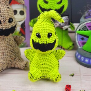 Puede incluir: Un juguete de peluche de crochet verde con forma de fantasma con una boca y ojos negros cosidos. El juguete est&aacute; sentado sobre una superficie de madera con otros juguetes de crochet y un tablero de juego en el fondo.