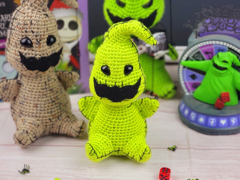 Puede incluir: Un juguete de peluche de crochet verde con forma de fantasma con una boca y ojos negros cosidos. El juguete est&aacute; sentado sobre una superficie de madera con otros juguetes de crochet y un tablero de juego en el fondo.