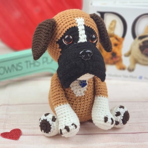Puede incluir: Un juguete de peluche de boxer de crochet marrón y blanco con ojos negros y nariz negra. El juguete lleva un collar azul con un dije de huella de pata. El juguete está sentado sobre una superficie de madera clara.