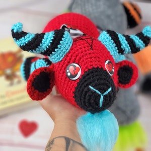 Peut inclure: Poup&eacute;e au crochet faite &agrave; la main avec une t&ecirc;te rouge, un visage noir et des cornes bleues. La poup&eacute;e a des yeux en forme de c&oelig;ur et une barbe bleue. Les cornes sont ray&eacute;es de noir et de bleu. Objet artisanal unique.