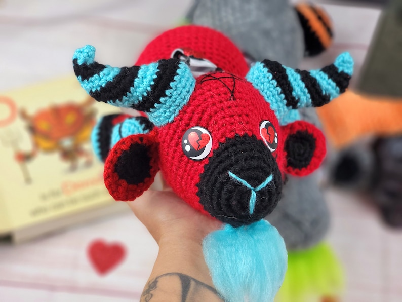 Peut inclure: Poup&eacute;e au crochet faite &agrave; la main avec une t&ecirc;te rouge, un visage noir et des cornes bleues. La poup&eacute;e a des yeux en forme de c&oelig;ur et une barbe bleue. Les cornes sont ray&eacute;es de noir et de bleu. Objet artisanal unique.