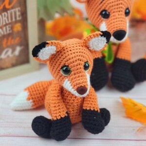 May include: Two crocheted fox plushies, one orange and white with black eyes and a green ribbon, the other orange and white with black eyes and a white tip on its tail. The plushies are sitting on a white surface with fall leaves.