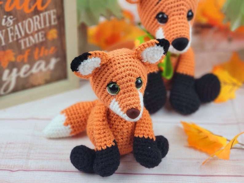 May include: Two crocheted fox plushies, one orange and white with black eyes and a green ribbon, the other orange and white with black eyes and a white tip on its tail. The plushies are sitting on a white surface with fall leaves.