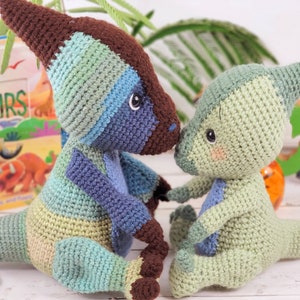 Pu&ograve; includere: Due giocattoli di dinosauro all'uncinetto, uno blu e verde e uno verde. I dinosauri sono seduti uno accanto all'altro e si guardano. I dinosauri sono fatti a mano e hanno una consistenza morbida e coccolabile.