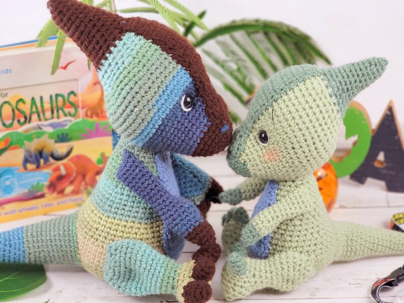 Pu&ograve; includere: Due giocattoli di dinosauro all'uncinetto, uno blu e verde e uno verde. I dinosauri sono seduti uno accanto all'altro e si guardano. I dinosauri sono fatti a mano e hanno una consistenza morbida e coccolabile.