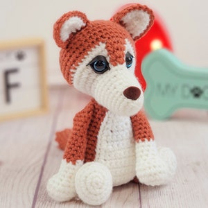 May include: A crocheted stuffed dog toy with brown and white fur. The dog has blue eyes and a brown nose. The toy is sitting on a wooden surface.