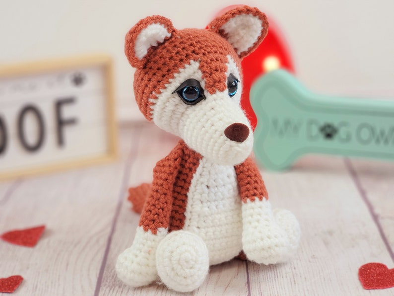 May include: A crocheted stuffed dog toy with brown and white fur. The dog has blue eyes and a brown nose. The toy is sitting on a wooden surface.