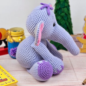 May include: A crocheted purple elephant toy with pink ears and a purple flower on its head. The elephant is sitting on a wooden surface.