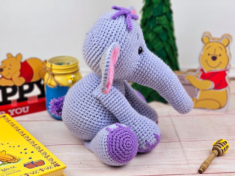 May include: A crocheted purple elephant toy with pink ears and a purple flower on its head. The elephant is sitting on a wooden surface.