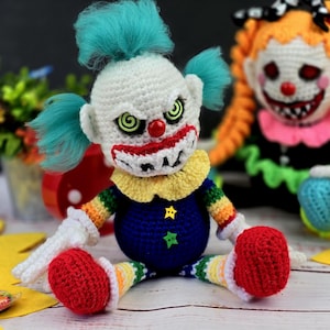 May include: Two crocheted clown dolls with creepy expressions. The doll on the left has a white face with a red nose, green eyes, and a wide, toothy grin. The doll on the right has an orange face with a red nose, black eyes, and a wide, toothy grin. Both dolls are wearing colourful clothing.
