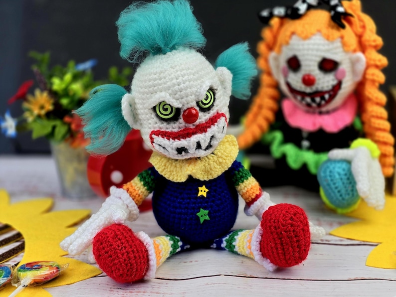 May include: Two crocheted clown dolls with creepy expressions. The doll on the left has a white face with a red nose, green eyes, and a wide, toothy grin. The doll on the right has an orange face with a red nose, black eyes, and a wide, toothy grin. Both dolls are wearing colourful clothing.