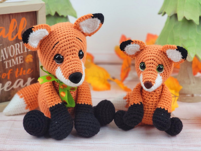 May include: Two crocheted fox plushies, one larger than the other. The larger fox is orange with black eyes and a white tail tip. The smaller fox is orange with black eyes and a white belly. Both foxes have black noses and black feet.