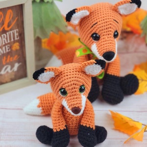 May include: Two crocheted fox plushies, one orange and one brown, with black eyes and noses. The orange fox is sitting with its tail curled around its legs. The brown fox is standing with its tail behind it. Both foxes have black feet.