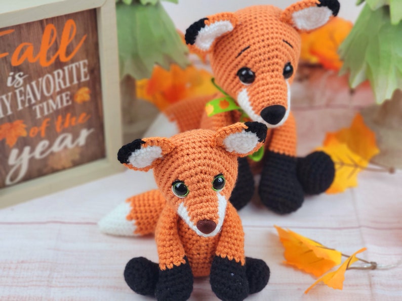 May include: Two crocheted fox plushies, one orange and one brown, with black eyes and noses. The orange fox is sitting with its tail curled around its legs. The brown fox is standing with its tail behind it. Both foxes have black feet.