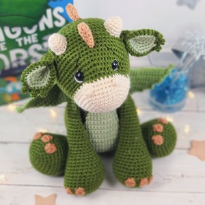 May include: A green crocheted dragon plush toy with a cream coloured snout and brown horns. The dragon is sitting with its wings out and has a sweet expression on its face.