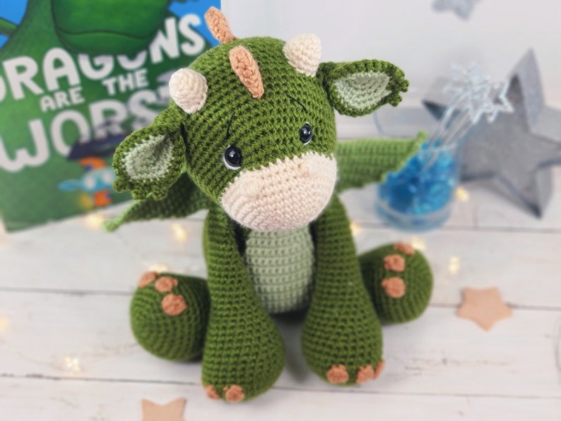 May include: A green crocheted dragon plush toy with a cream coloured snout and brown horns. The dragon is sitting with its wings out and has a sweet expression on its face.