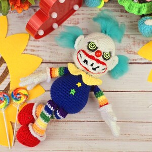 May include: A crocheted clown doll with a blue and white striped outfit, red shoes, and a creepy smile. The doll is lying on a wooden surface with a sunflower and lollipops nearby.