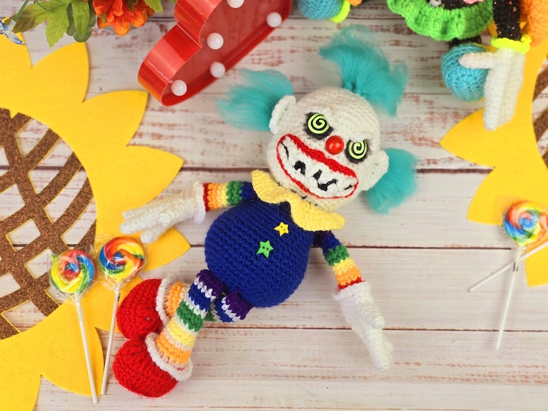 May include: A crocheted clown doll with a blue and white striped outfit, red shoes, and a creepy smile. The doll is lying on a wooden surface with a sunflower and lollipops nearby.
