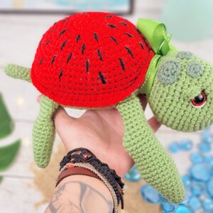 Puede incluir: Un juguete de tortuga de crochet con una c&aacute;scara de sand&iacute;a. La tortuga es verde con ojos negros y un lazo verde.