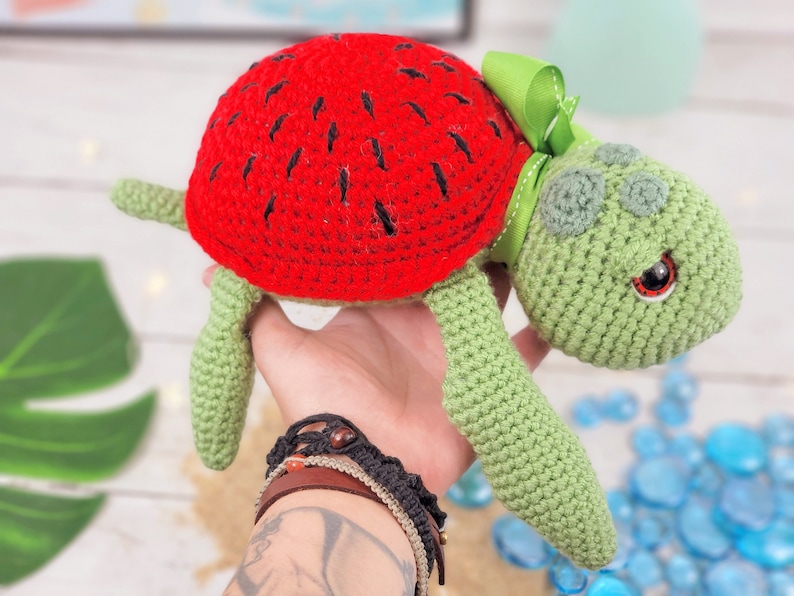 Puede incluir: Un juguete de tortuga de crochet con una c&aacute;scara de sand&iacute;a. La tortuga es verde con ojos negros y un lazo verde.