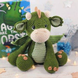 May include: A green crocheted dragon toy with brown feet and a light brown snout. The dragon is sitting on a white surface with a book titled "Dragons are the Worst" in the background.