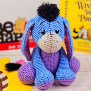 May include: A crocheted blue and purple Eeyore plush toy with a black fur mane. The toy is sitting on a yellow surface with a brown and gold sunflower design.