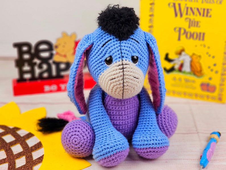 May include: A crocheted blue and purple Eeyore plush toy with a black fur mane. The toy is sitting on a yellow surface with a brown and gold sunflower design.