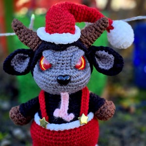 Puede incluir: Una cabra de crochet gris y negro con un gorro de Pap&aacute; Noel rojo con ribete blanco y un mono rojo con estrellas doradas. La cabra tiene ojos rojos y una lengua rosa.