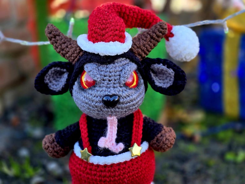 Puede incluir: Una cabra de crochet gris y negro con un gorro de Pap&aacute; Noel rojo con ribete blanco y un mono rojo con estrellas doradas. La cabra tiene ojos rojos y una lengua rosa.