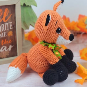 May include: A crocheted orange fox with black feet and a white tipped tail. The fox is wearing a green and orange ribbon around its neck. The fox is sitting on a white surface with fall leaves in the background.
