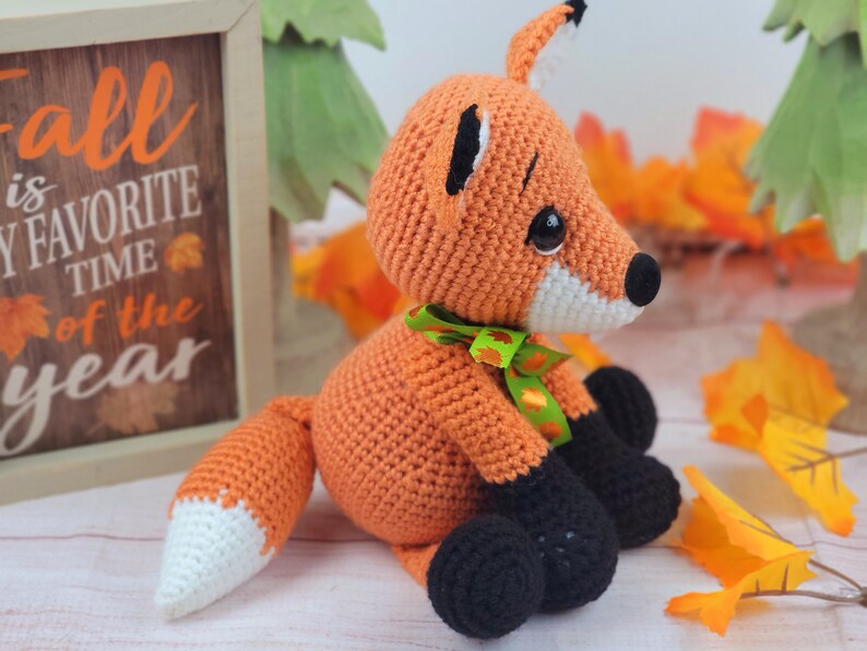 May include: A crocheted orange fox with black feet and a white tipped tail. The fox is wearing a green and orange ribbon around its neck. The fox is sitting on a white surface with fall leaves in the background.