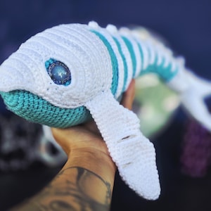 Può includere: Un peluche a forma di balena all'uncinetto fatto a mano in bianco e verde acqua. La balena presenta un design dettagliato con un corpo bianco, accenti verde acqua e un dettaglio con gli occhi stellati. Il peluche è un giocattolo morbido e coccoloso.