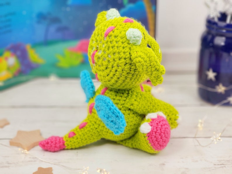 May include: A green crocheted dragon with pink and blue accents. The dragon has a small, curled tail and is sitting on a white surface.