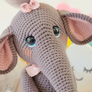 May include: A gray crocheted elephant toy with pink ears and a pink bow. The elephant has blue eyes and a stitched smile.