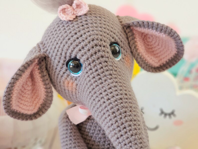 May include: A gray crocheted elephant toy with pink ears and a pink bow. The elephant has blue eyes and a stitched smile.