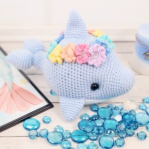 K&ouml;nnte beinhalten: Ein hellblauer geh&auml;kelter Delfin mit einer Regenbogenblumenkrone. Der Delfin sitzt auf einer wei&szlig;en Oberfl&auml;che mit blauen Glassteinen.