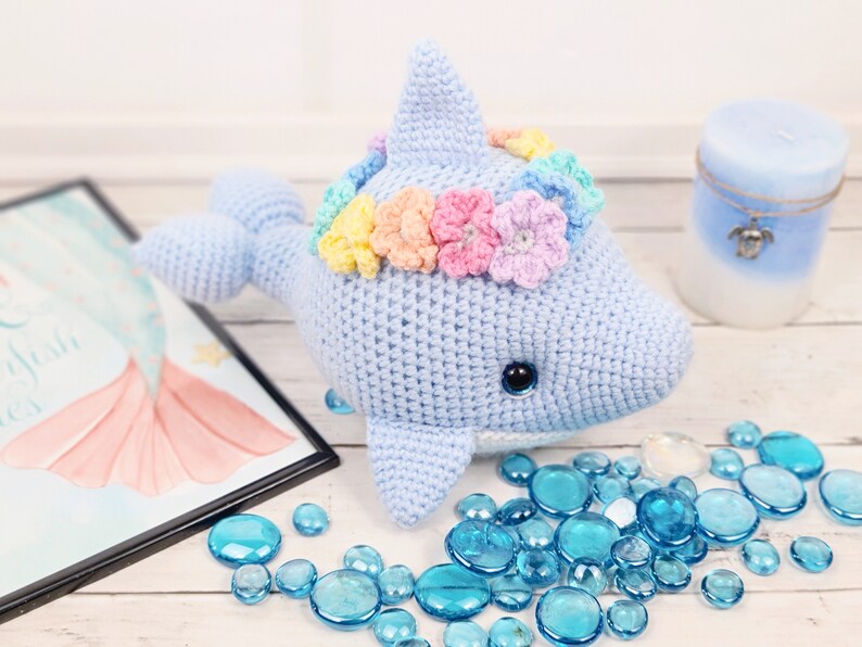 K&ouml;nnte beinhalten: Ein hellblauer geh&auml;kelter Delfin mit einer Regenbogenblumenkrone. Der Delfin sitzt auf einer wei&szlig;en Oberfl&auml;che mit blauen Glassteinen.