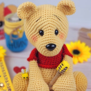 K&ouml;nnte beinhalten: Ein geh&auml;keltes Winnie Puuh-B&auml;ren-Spielzeug mit einem roten Shirt und einem Honigstock. Der B&auml;r ist gelb und braun und sitzt auf einer wei&szlig;en Oberfl&auml;che.
