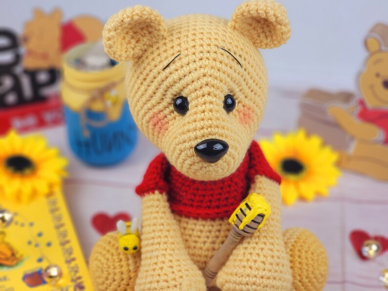 K&ouml;nnte beinhalten: Ein geh&auml;keltes Winnie Puuh-B&auml;ren-Spielzeug mit einem roten Shirt und einem Honigstock. Der B&auml;r ist gelb und braun und sitzt auf einer wei&szlig;en Oberfl&auml;che.