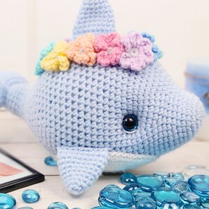 K&ouml;nnte beinhalten: Ein hellblauer geh&auml;kelter Delfin mit einer Regenbogenblumenkrone. Der Delfin hat einen wei&szlig;en Bauch und schwarze Augen. Der Delfin sitzt auf einer wei&szlig;en Oberfl&auml;che mit blauen Glassteinen.