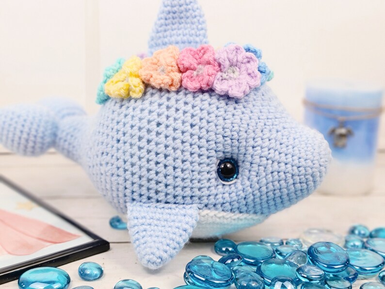 K&ouml;nnte beinhalten: Ein hellblauer geh&auml;kelter Delfin mit einer Regenbogenblumenkrone. Der Delfin hat einen wei&szlig;en Bauch und schwarze Augen. Der Delfin sitzt auf einer wei&szlig;en Oberfl&auml;che mit blauen Glassteinen.