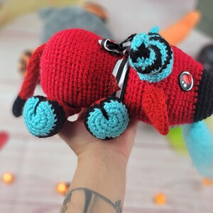 Peut inclure: Un jouet rouge fait &agrave; la main au crochet avec des accents noirs et turquoise. Le jouet pr&eacute;sente un museau noir, des pieds turquoise et un n&oelig;ud d&eacute;coratif &agrave; rayures noires et blanches. Un bouton en forme de c&oelig;ur est sur le c&ocirc;t&eacute;.