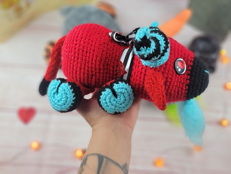 Peut inclure: Un jouet rouge fait &agrave; la main au crochet avec des accents noirs et turquoise. Le jouet pr&eacute;sente un museau noir, des pieds turquoise et un n&oelig;ud d&eacute;coratif &agrave; rayures noires et blanches. Un bouton en forme de c&oelig;ur est sur le c&ocirc;t&eacute;.