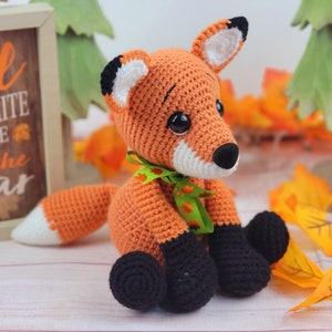 May include: A crocheted orange fox with black eyes, a white nose, and black feet. The fox is wearing a green ribbon with orange and white leaves. The fox is sitting on a white wooden surface with fall leaves in the background.