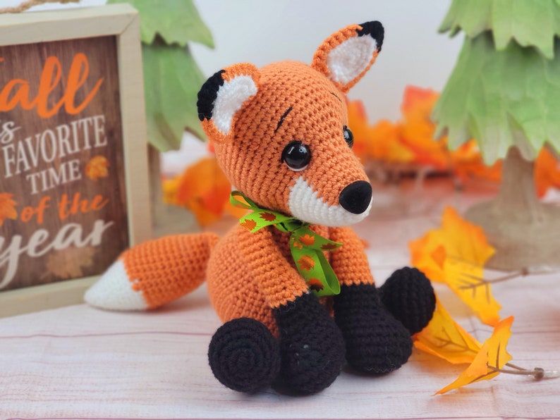 May include: A crocheted orange fox with black eyes, a white nose, and black feet. The fox is wearing a green ribbon with orange and white leaves. The fox is sitting on a white wooden surface with fall leaves in the background.