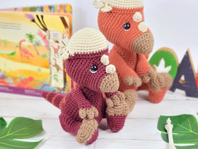 Pu&ograve; includere: Due giocattoli di dinosauro all'uncinetto, uno rosso con la testa color crema e uno arancione. I giocattoli sono seduti su una superficie bianca con foglie verdi.