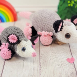 May include: Two crocheted opossum plushies, one grey and white and one white with pink accents. The plushies have black eyes and pink noses.