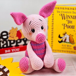 May include: A pink crocheted pig stuffed animal with a striped pink and purple shirt. The pig is sitting on a wooden surface with a book titled "The Complete Tales of Winnie the Pooh" in the background.
