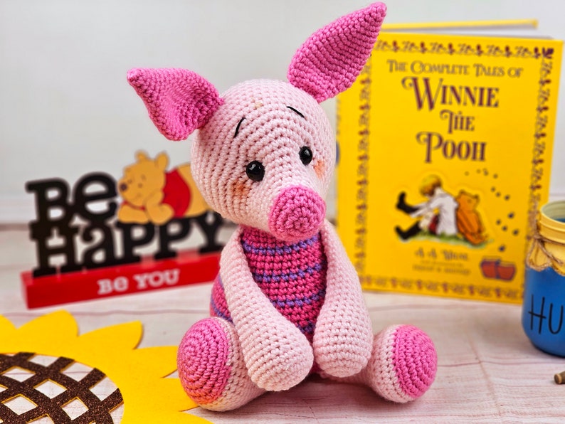 May include: A pink crocheted pig stuffed animal with a striped pink and purple shirt. The pig is sitting on a wooden surface with a book titled "The Complete Tales of Winnie the Pooh" in the background.