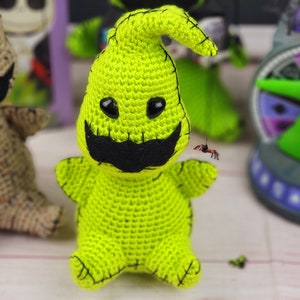 Puede incluir: Un juguete de amigurumi de crochet verde con forma de fantasma con una boca cosida negra y un bigote negro. El juguete tiene ojos negros y un gorro de crochet verde.